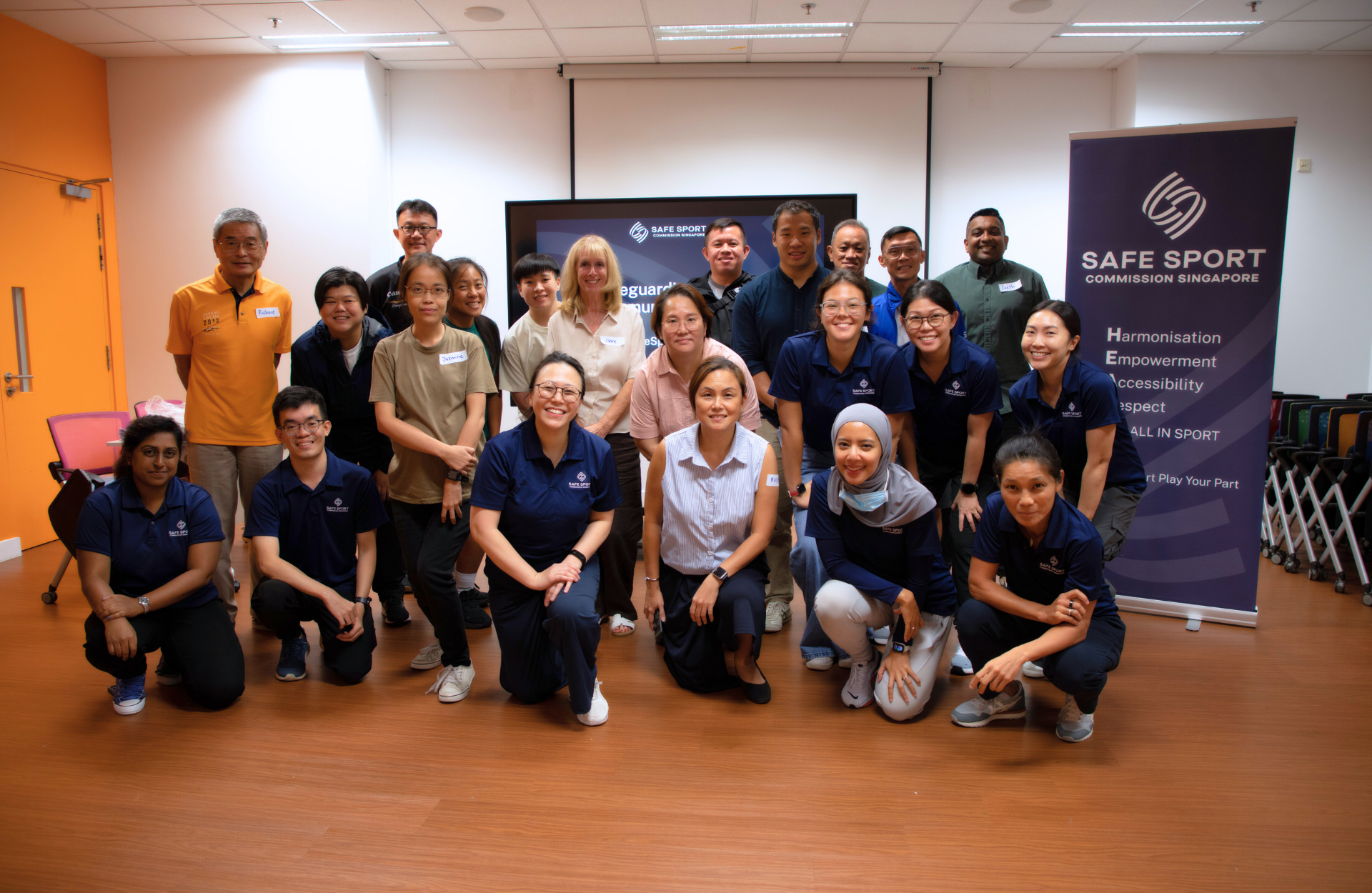 Group of people posing indoors with SAFE SPORT Commission Singapore banner and TV screen in the background.